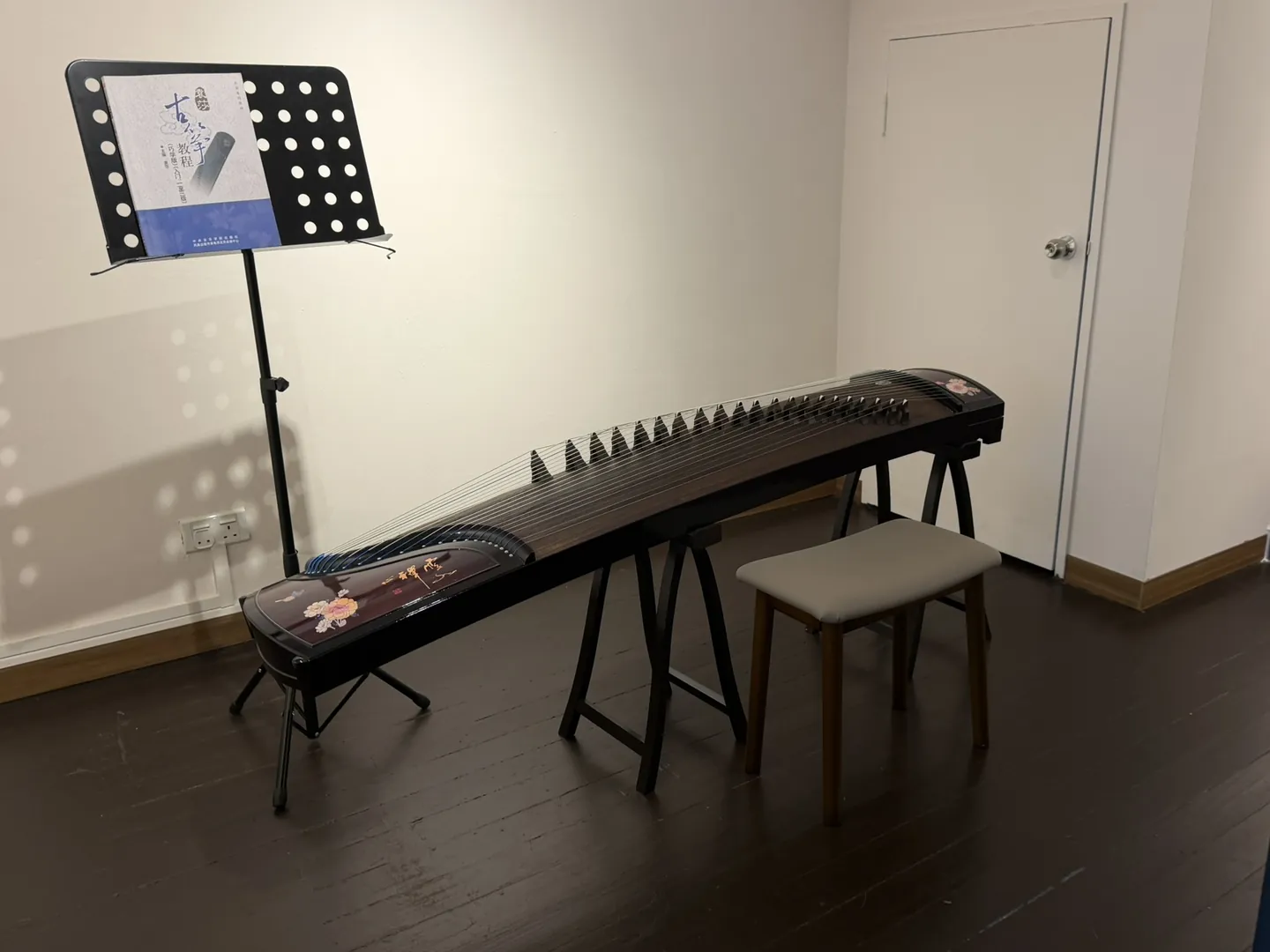 A guzheng on its stand in a teaching room, with a stool in front of it and a music stand holding a guzheng tutor book
