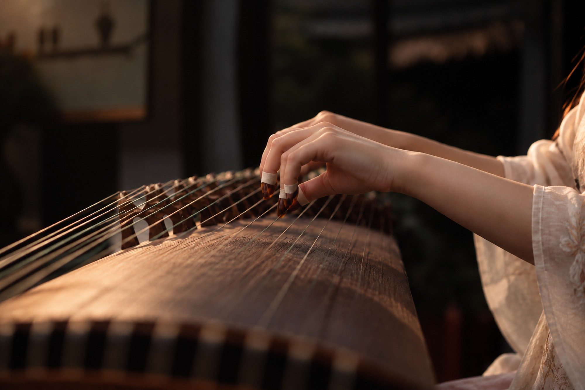 Close-up of a guzheng player’s hand, with fingerpicks, plucking the strings in warm, low light